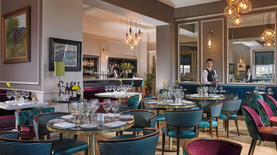 Elegant hotel restaurant with stylish decor, plush seating, ambient lighting; waiter sets inviting tables with fine dining settings.
