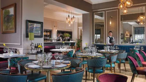 Elegant hotel restaurant with stylish decor, plush seating, ambient lighting; waiter sets inviting tables with fine dining settings.