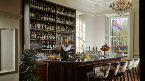 Elegant hotel bar with floral chairs, a bartender mixing drinks, and natural light from a large window.