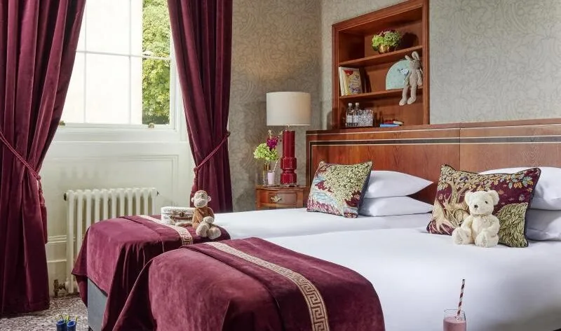 Hotel room with two single beds, purple decor, plush toys on each bed, a window with curtains, a bookshelf, and a table with a drink, book, and crayons in the foreground.