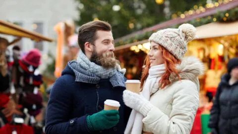Smiling couple in warm clothing enjoying hot drinks at a festive Christmas market, surrounded by twinkling lights.