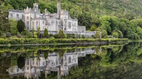 A large stone castle stands by a calm lake, surrounded by green trees and hills, with its reflection visible in the water.