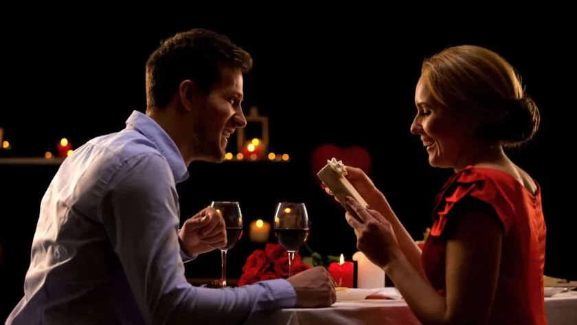 A couple enjoying a romantic candlelit dinner with red wine and a gift, surrounded by soft glowing candles.