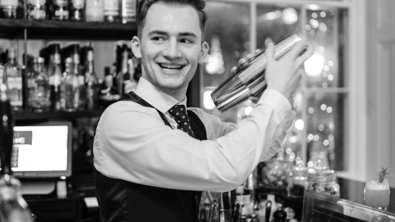 Smiling bartender preparing cocktails in stylish hotel bar with illuminated shelves and window view.