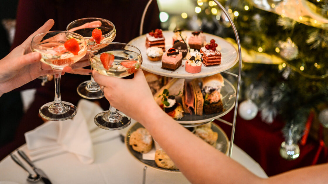 Guests enjoying afternoon tea, champagne, and festive treats by a decorated Christmas tree in elegant hotel setting.