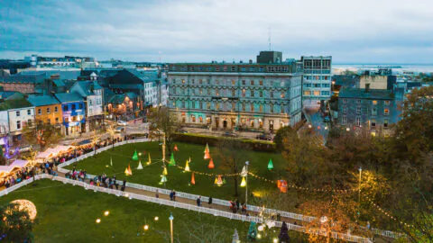 Aerial view of festive city square with colourful lights, bustling winter market, and historic hotel backdrop at twilight.