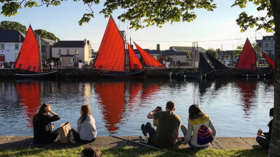 Guests enjoy a sunny day by the river, admiring traditional boats with vibrant red sails, perfect for a relaxing getaway.