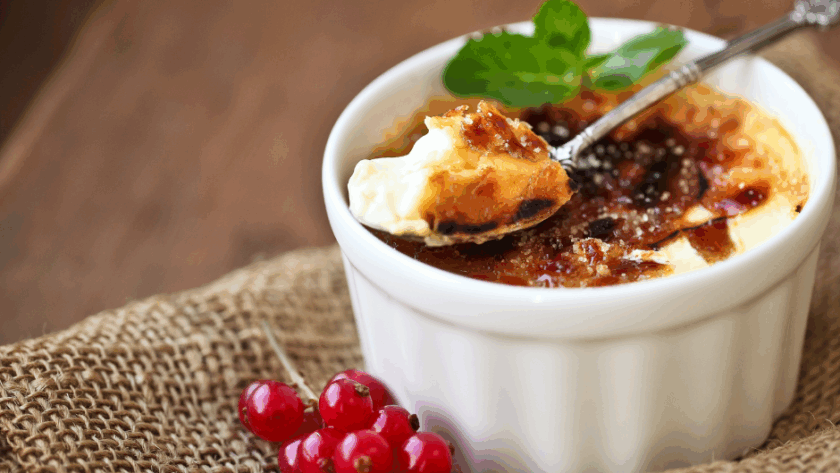 A ramekin of cr&egrave;me br&ucirc;l&eacute;e with caramelized sugar top, garnished with mint and red currants, sits on burlap with a spoonful taken out.