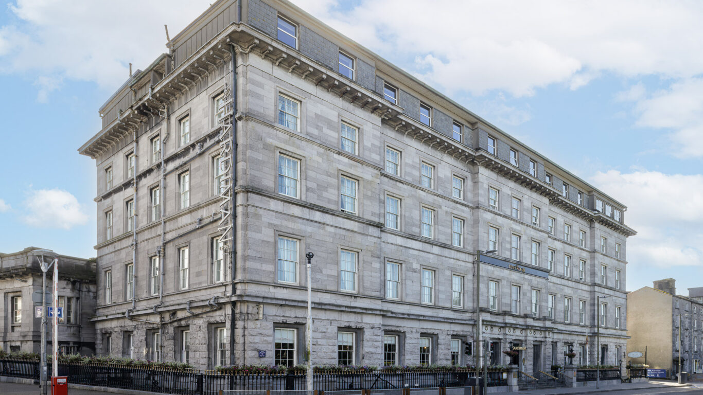 Historic grey stone hotel facade against blue sky, corner view; charming urban retreat with traditional elegance.