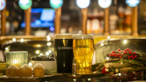 A pint of Guinness and a pint of Connemara beer on a bar counter with desserts, olives, and festive decorations in the background.
