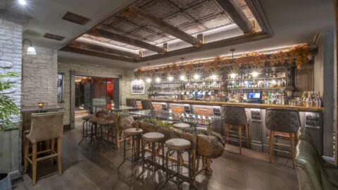 Cosy hotel bar with rustic decor, plush stools, wood ceiling, inviting ambience, wide drink selection, perfect for relaxation.