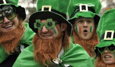 Four people dressed as leprechauns with green hats, orange beards, capes, and face paint, smiling and celebrating outdoors.