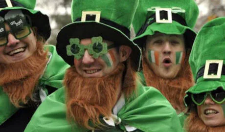 Four people dressed as leprechauns with green hats, orange beards, capes, and face paint, smiling and celebrating outdoors.