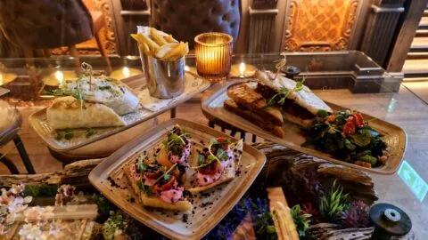 Three plates of food on a glass table: a wrap with fries, sandwiches with salad, and toast topped with pink spread and black sesame seeds. A lit candle sits in the background.