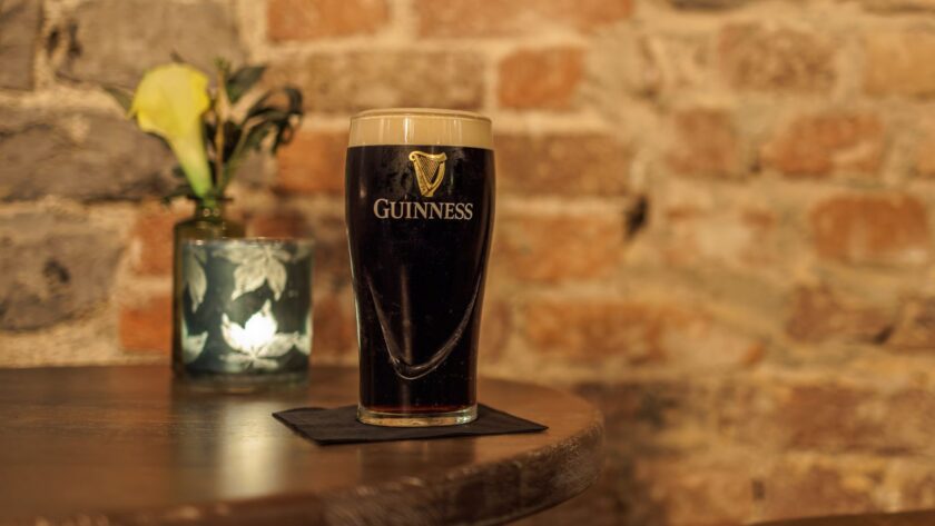 A pint glass of Guinness stout on a coaster sits on a wooden table, with a flower vase and a candle in the background against a brick wall.