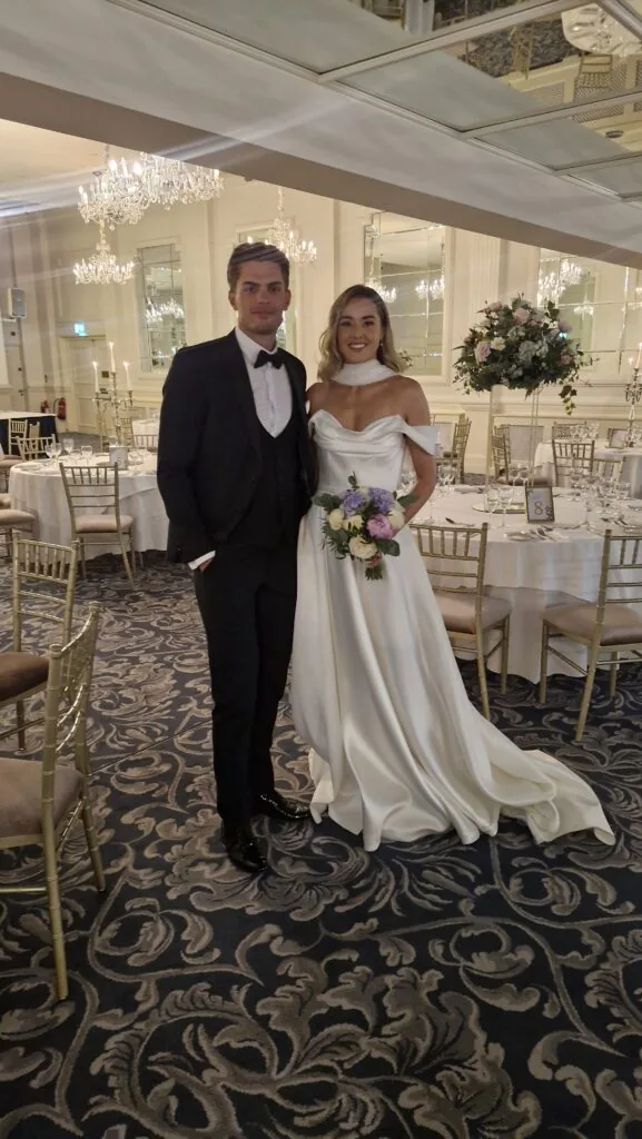 A bride in a white dress and groom in a black tuxedo pose together in an elegant, chandelier-lit wedding reception hall.