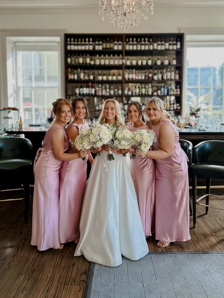 Five women in formal dresses, including a bride in white and four bridesmaids in pink, pose together holding bouquets in front of a bar with shelves of bottles.
