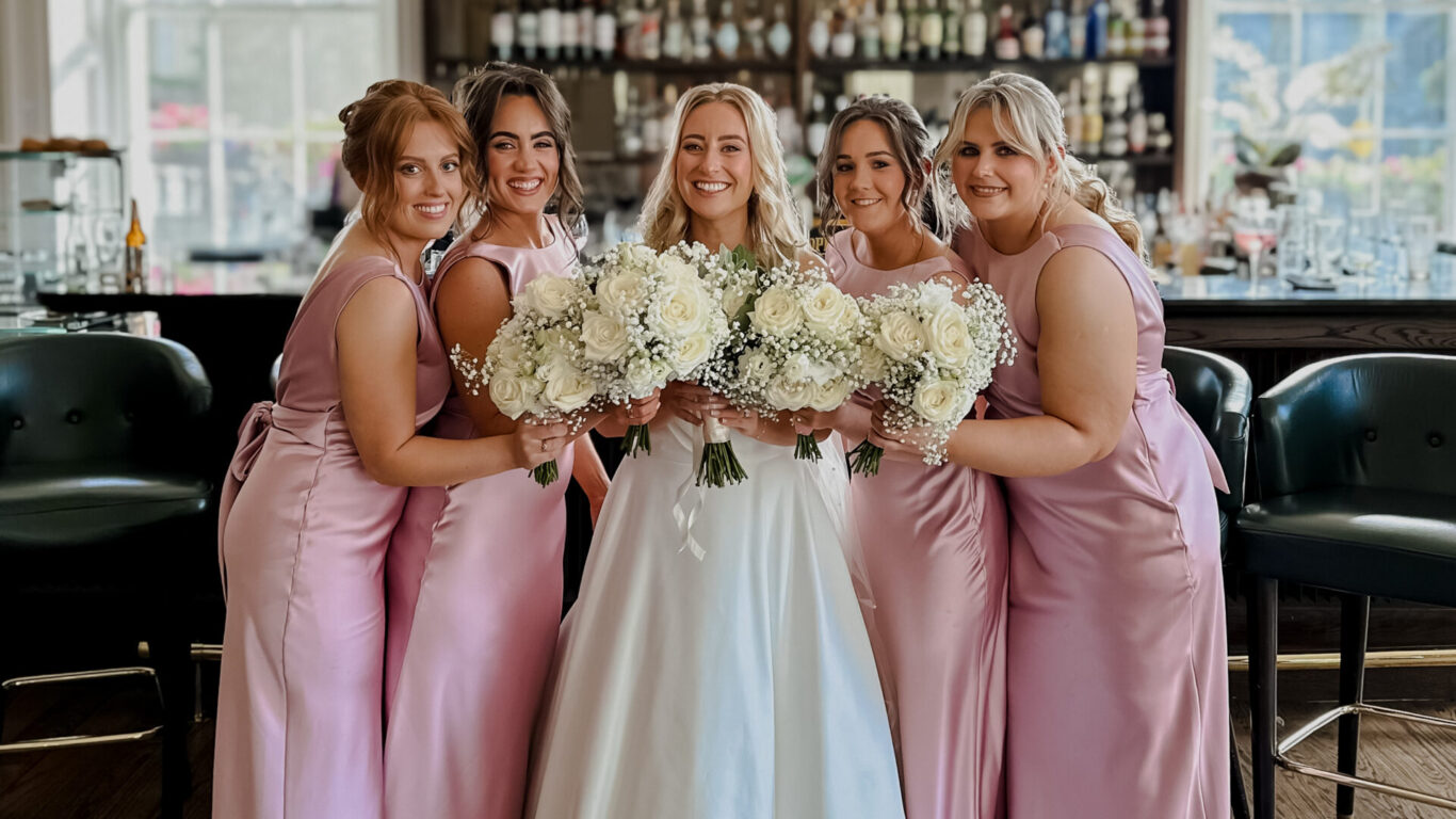 Five women in formal dresses, including a bride in white and four bridesmaids in pink, pose together holding bouquets in front of a bar with shelves of bottles.