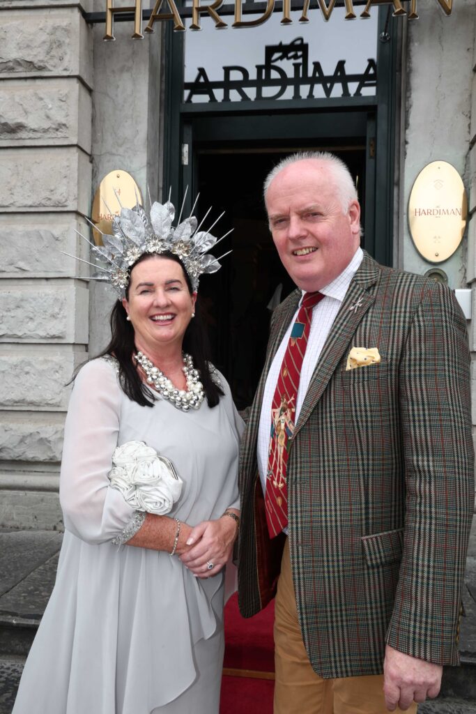 A woman in a light grey dress and ornate silver headpiece stands next to a man in a plaid jacket and red tie, both smiling outside a building with 