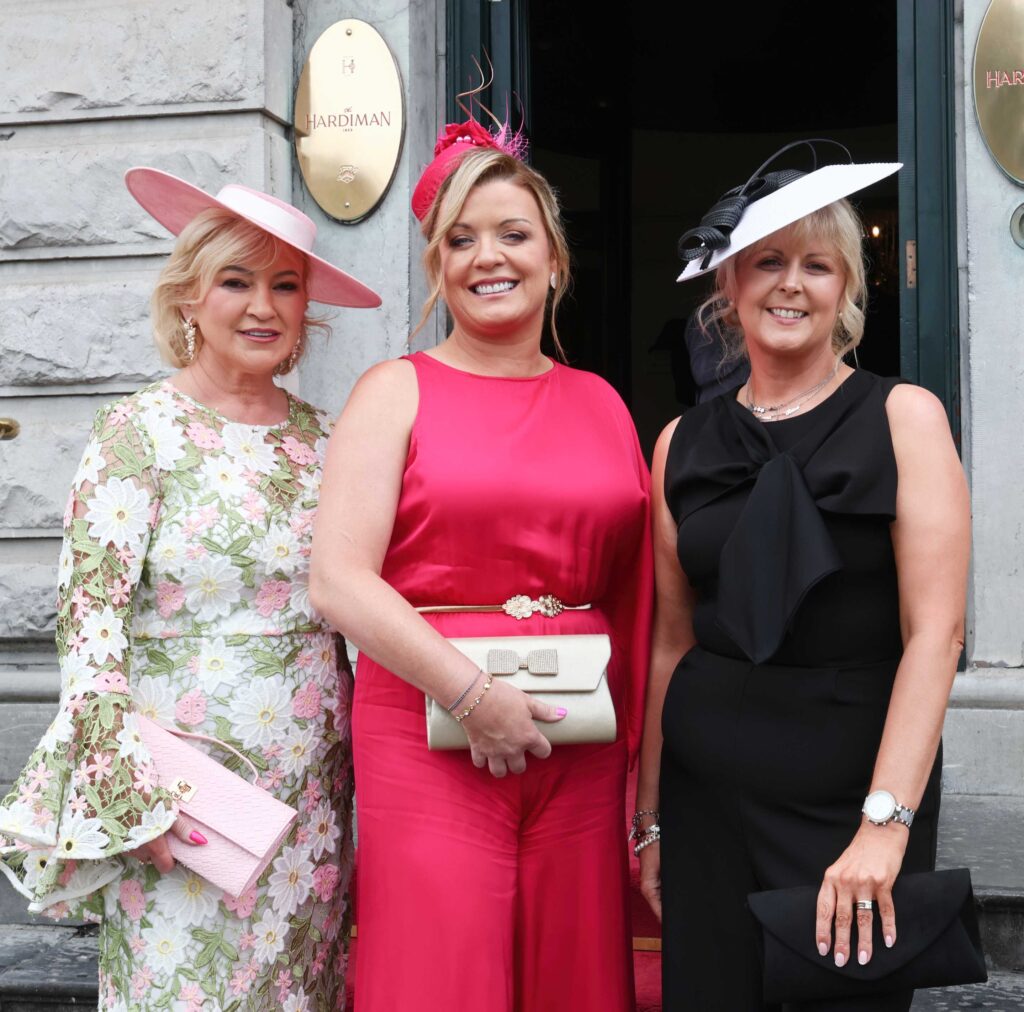 Three women in formal attire and hats pose and smile outside a building with stone walls and a sign reading 