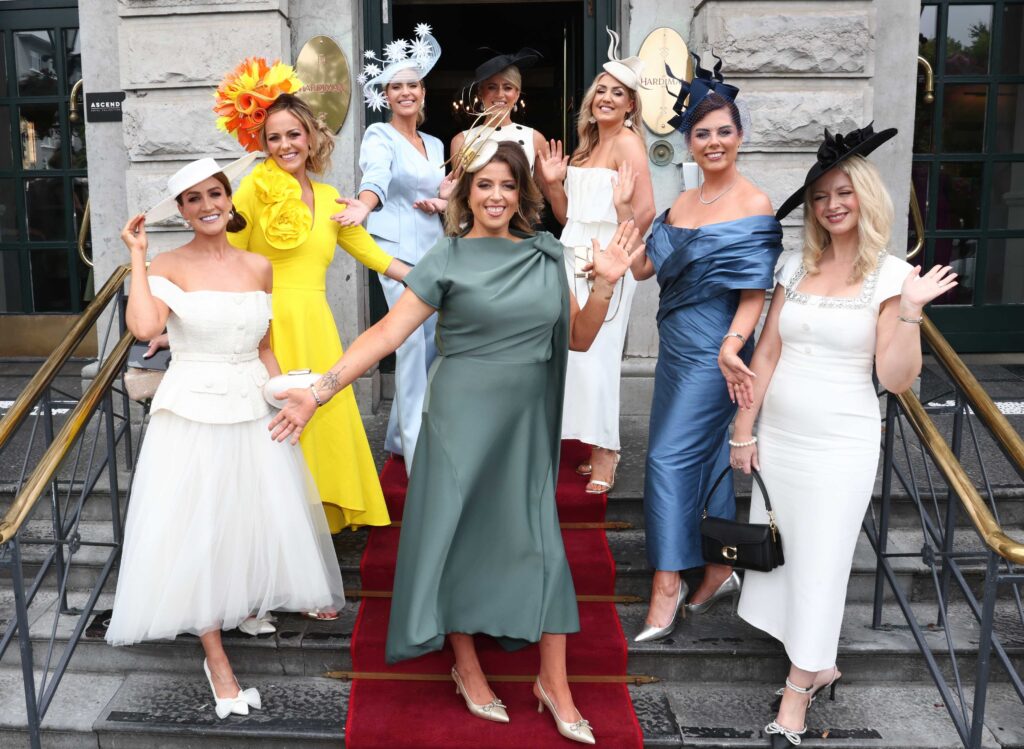 Eight women dressed in colorful formal outfits and hats pose and smile on a red carpeted staircase outside a building.