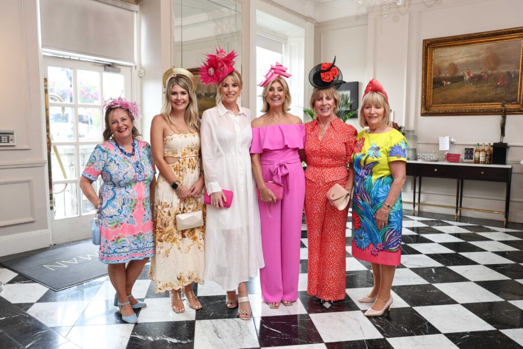 Six women dressed in bright, formal outfits and hats stand together and pose for a photo in a room with black and white checkered flooring.
