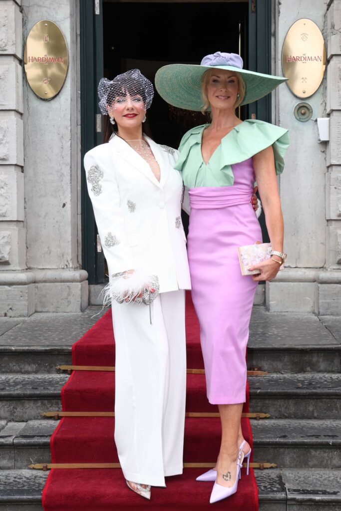 Two women pose on a red carpet; one wears a white suit with a fascinator, the other wears a lavender dress with a wide-brimmed hat and green accents.