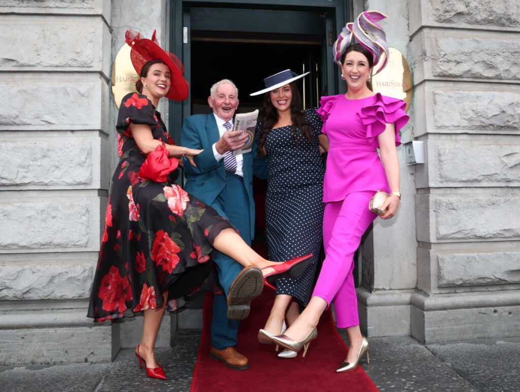 Three women in colorful, formal outfits and hats pose with an older man in a suit on a red carpet outside a stone building, all smiling and lifting one leg playfully.