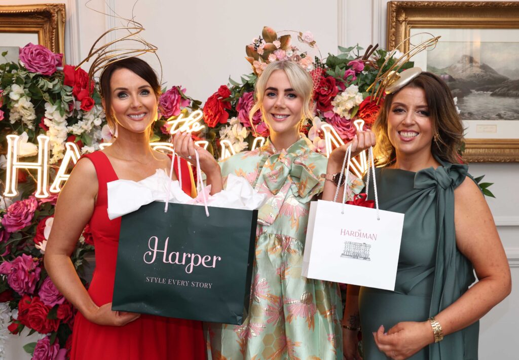 Three women in formal dresses and decorative headpieces hold gift bags, standing in front of a floral display and illuminated 