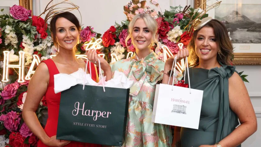Three women in formal dresses and decorative headpieces hold gift bags, standing in front of a floral display and illuminated 