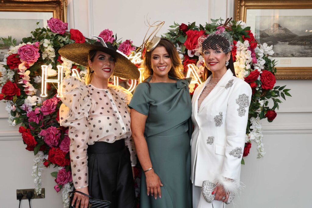 Three women in formal attire and hats pose together in front of a flower wall with illuminated lettering at an indoor event.