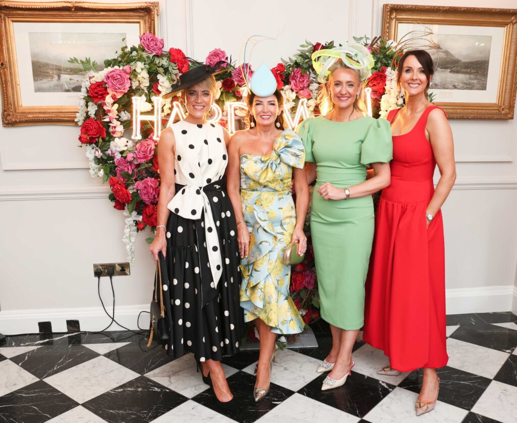 Four women dressed in colorful formal outfits and hats stand on a black-and-white checkered floor in front of a flower heart and framed pictures on a wall.