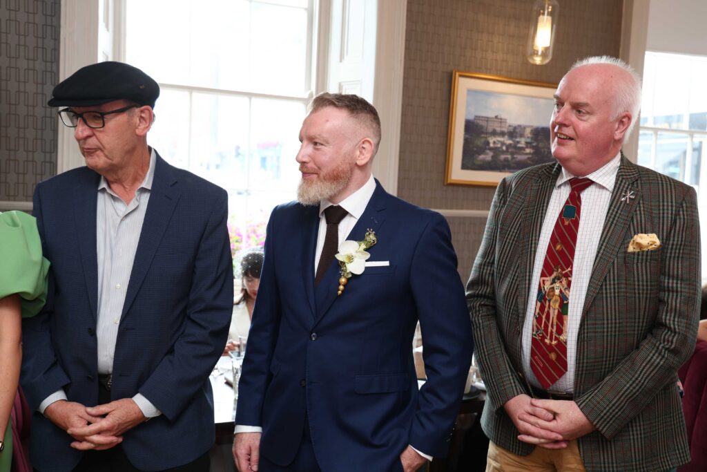 Three men in suits stand indoors at a formal event; one wears a beret, another has a boutonniere, and the third wears a patterned blazer and festive tie.