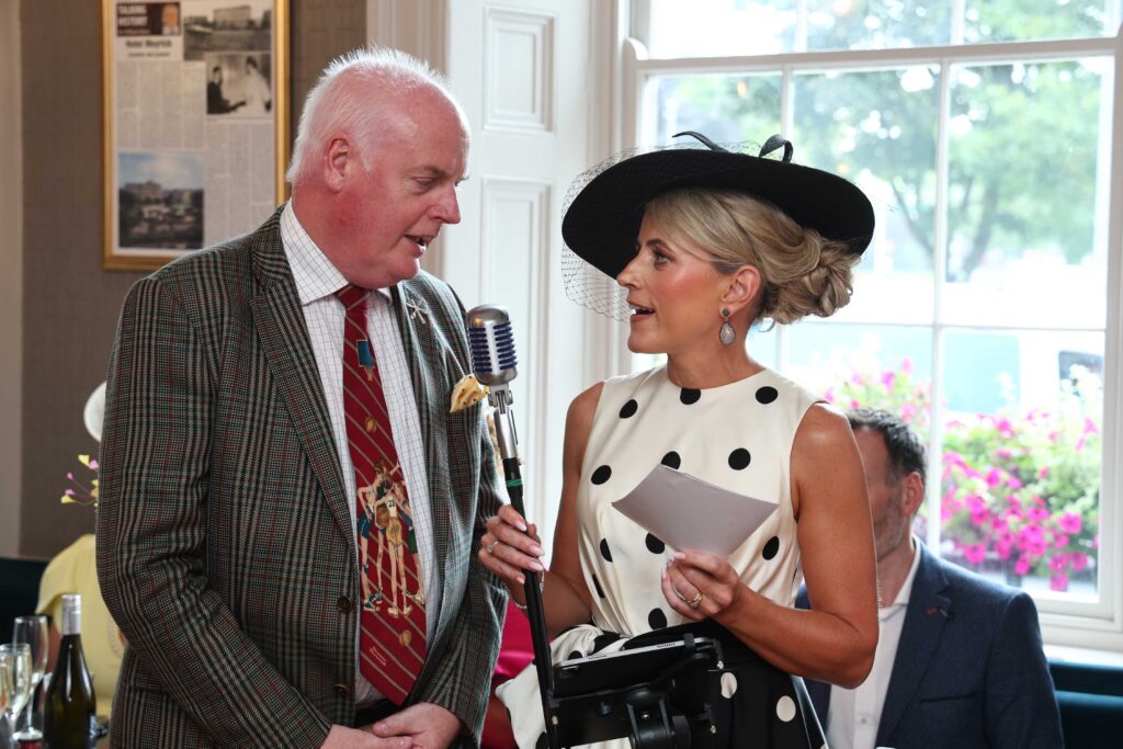 A man and a woman dressed in formal attire speak into a vintage microphone by a window; the woman holds a card and wears a large black hat with netting.