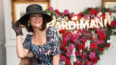 A woman in a patterned dress and black hat poses indoors in front of a neon