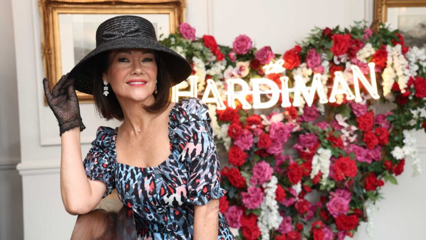A woman in a patterned dress and black hat poses indoors in front of a neon 