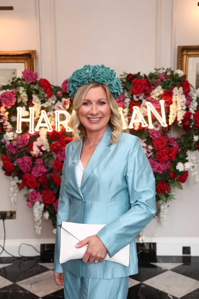 A woman in a light blue suit and teal floral headpiece stands indoors, holding a white clutch, with a floral heart and illuminated 