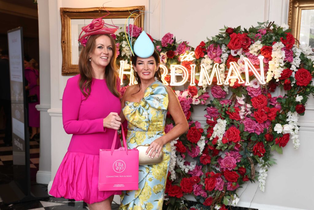 Two women in colorful dresses and hats pose in front of a neon sign and floral display, one holding a pink shopping bag.