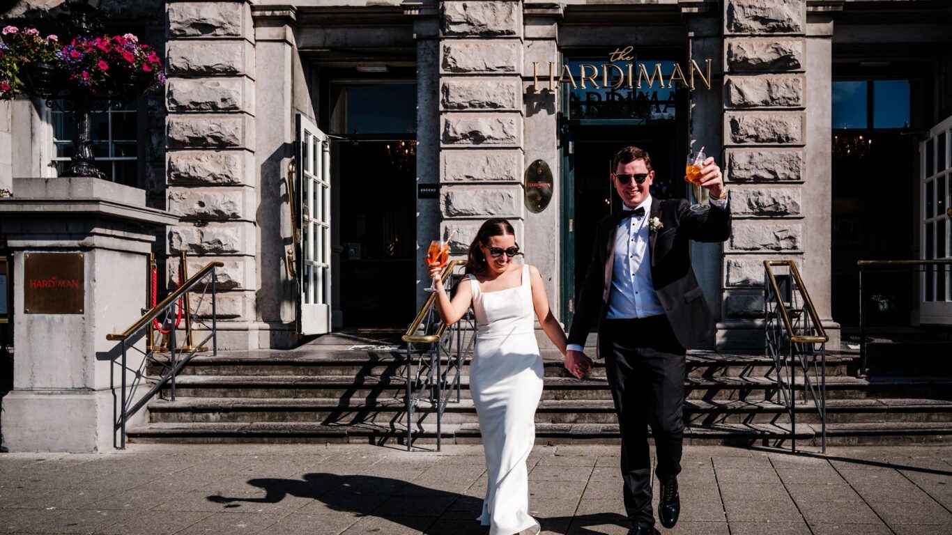 A bride and groom in formal attire hold hands and raise drinks while walking outside The Hardiman hotel on a sunny day.