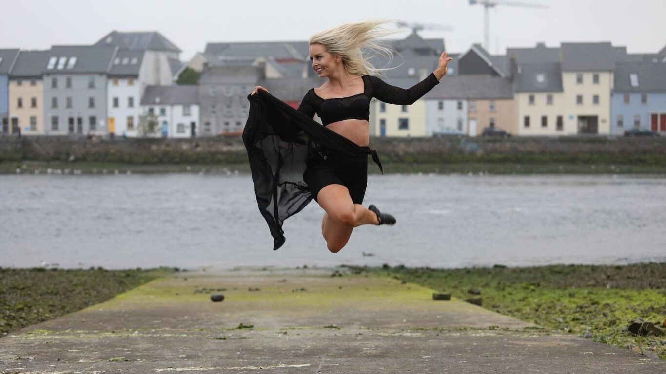 A woman in a black outfit jumps mid-air with an arm extended, holding sheer fabric, near a waterfront with houses and cranes in the background.