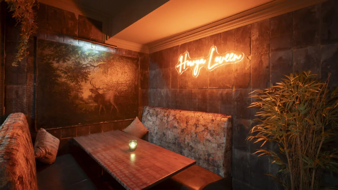 A cozy booth with a wooden table, cushioned seating, a plant, dim lighting, and a neon sign reading