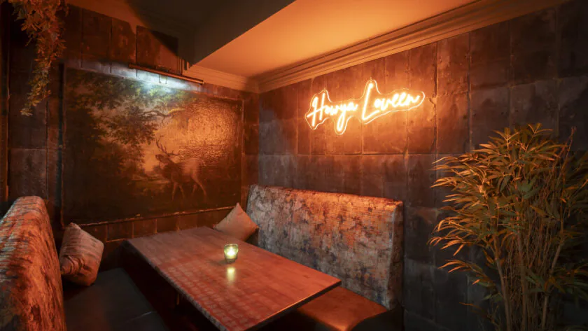 A cozy booth with a wooden table, cushioned seating, a plant, dim lighting, and a neon sign reading 
