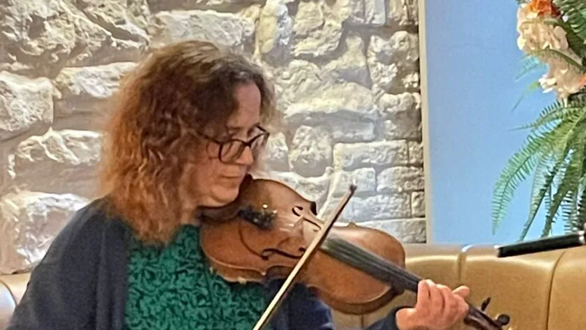 A woman with curly hair and glasses plays a violin while seated on a brown sofa in front of a stone wall and floral arrangement.