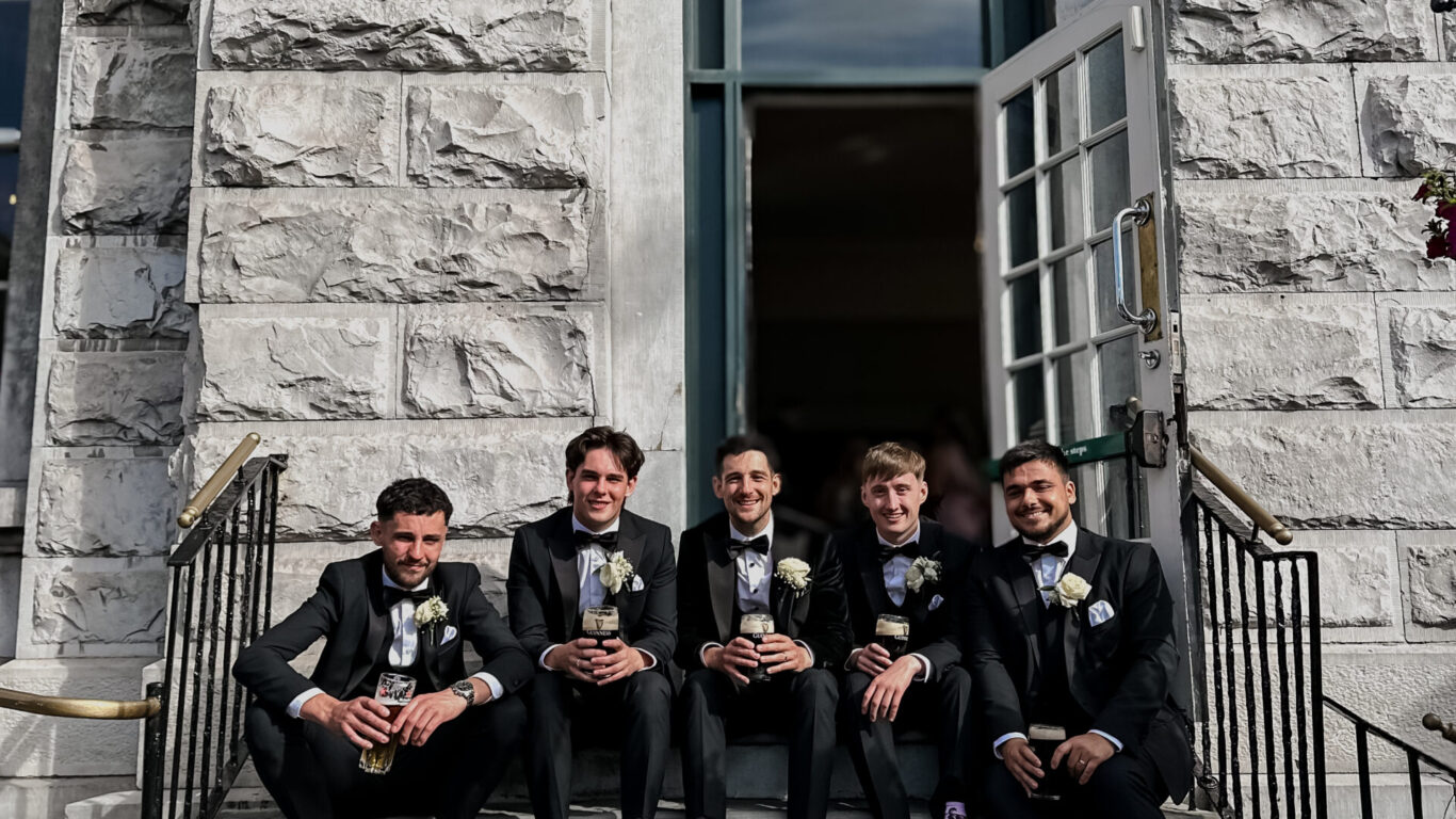 Five men in suits sit on stone steps outside a building with a large window and open door, posing for a group photo.