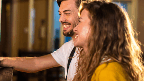 A man and a woman sit side by side at a bar, smiling and laughing, with warm indoor lighting in the background.