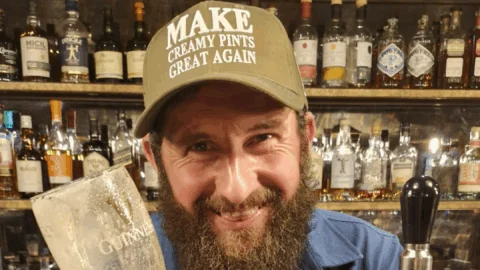 A bearded man in a blue shirt stands behind a bar holding an empty Guinness glass and smiling. He wears a cap that reads, 