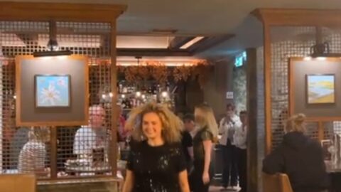A woman in black walks confidently through a restaurant as people dine and watch; someone in the foreground films her with a smartphone.