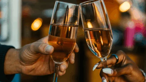 Two people clinking champagne glasses together in a celebratory toast, with a blurred background and warm lighting.