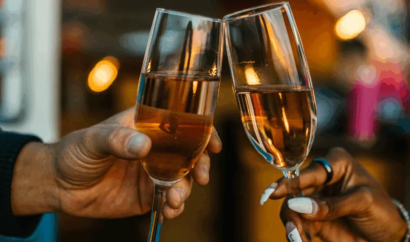 Two people clinking champagne glasses together in a celebratory toast, with a blurred background and warm lighting.