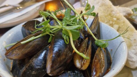 A bowl of cooked mussels garnished with microgreens, served with slices of bread on a plate, with a candle and tableware in the background.